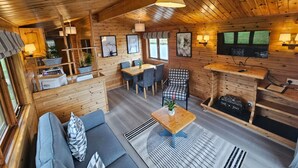 L’intérieur chaleureux d’un chalet avec un plafond en bois, un coin repas, un espace salon comprenant un canapé et des fauteuils, et un téléviseur fixé au mur.