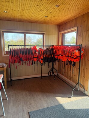 A room with wooden walls and ceiling, two windows with a view of trees, a rack with red life jackets, and a gray carpet.