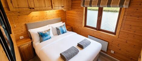 A wooden cabin bedroom with a bed, pillows, a window, and a view of a body of water.