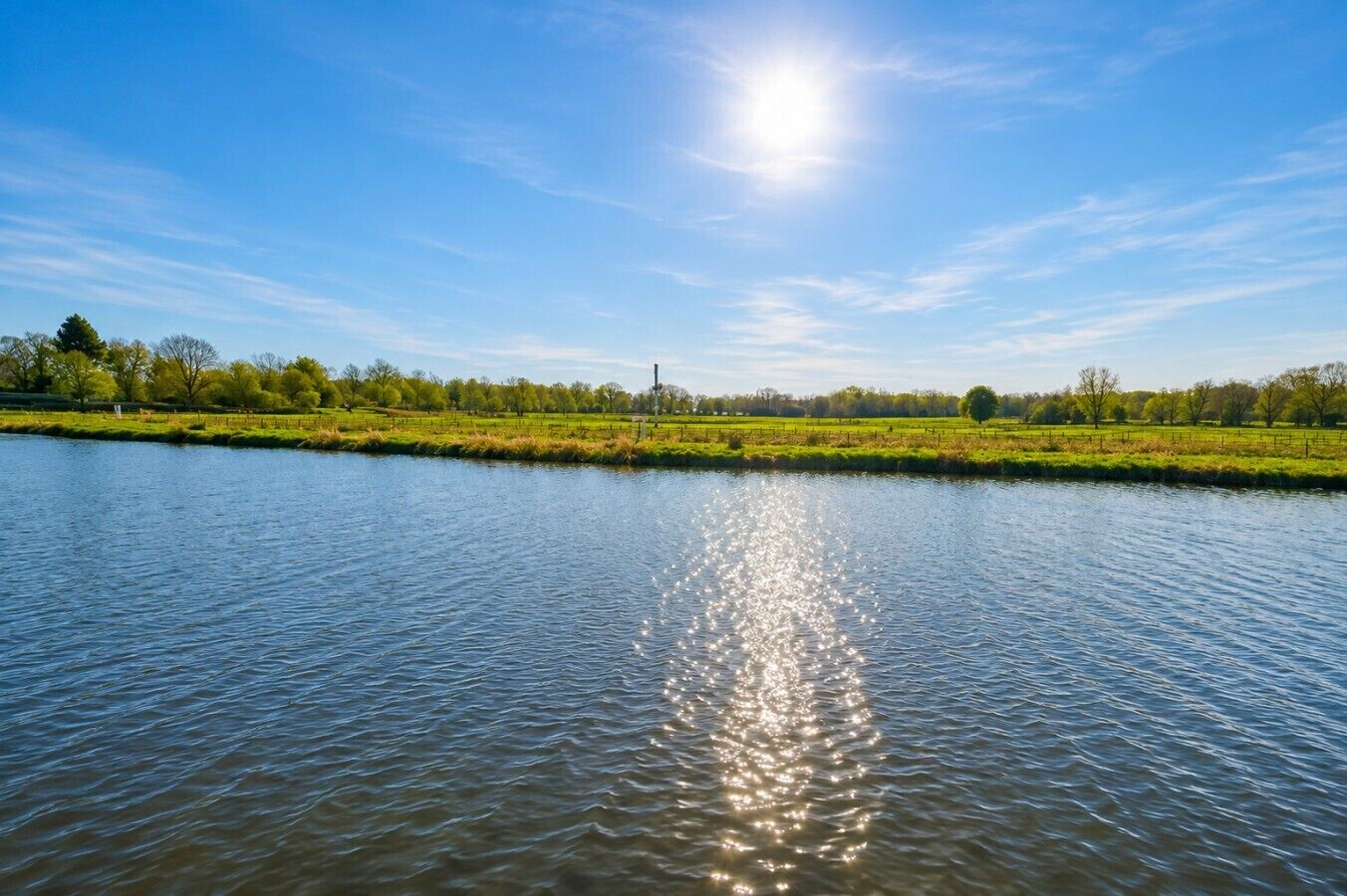 Un río tranquilo que refleja la luz del sol, rodeado de campos verdes y árboles bajo un cielo despejado.