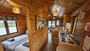 A cabin interior with wooden walls, a kitchenette, two beds with pillows, and a window overlooking a natural landscape.