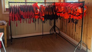 A room with wooden walls and ceiling, two windows with a view of trees, a rack with red life jackets, and a gray carpet.