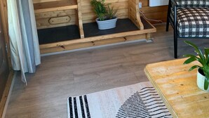Une pièce chaleureuse avec des murs en bois, une cheminée, deux lampes fixées au mur, un fauteuil à carreaux et une petite table sur laquelle est posée une plante.