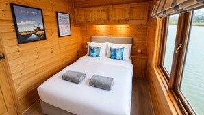 A wooden cabin bedroom with a bed, pillows, a nightstand, and a window with a view of water. - Wroxham Broads Lodge Houseboat (Stalham)