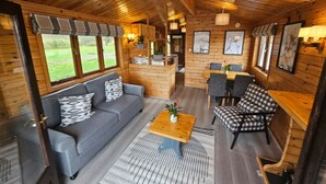 A cozy cabin interior with a sofa, armchair, and dining area, featuring wooden walls and large windows. - Wroxham Broads Lodge Houseboat (Stalham)