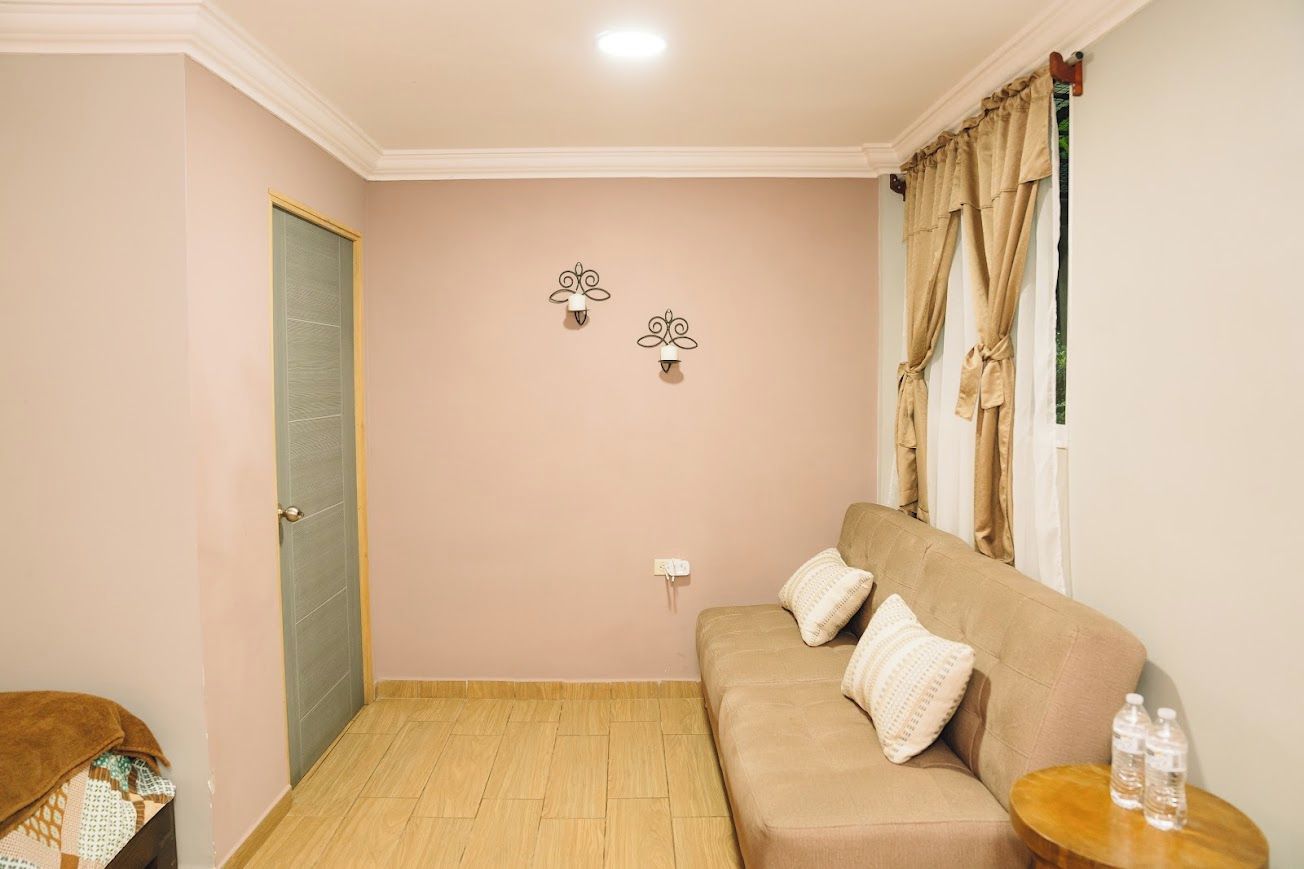 Una habitación con un sofá color beige, dos luces fijadas a la pared, una puerta y una ventana con cortinas.
