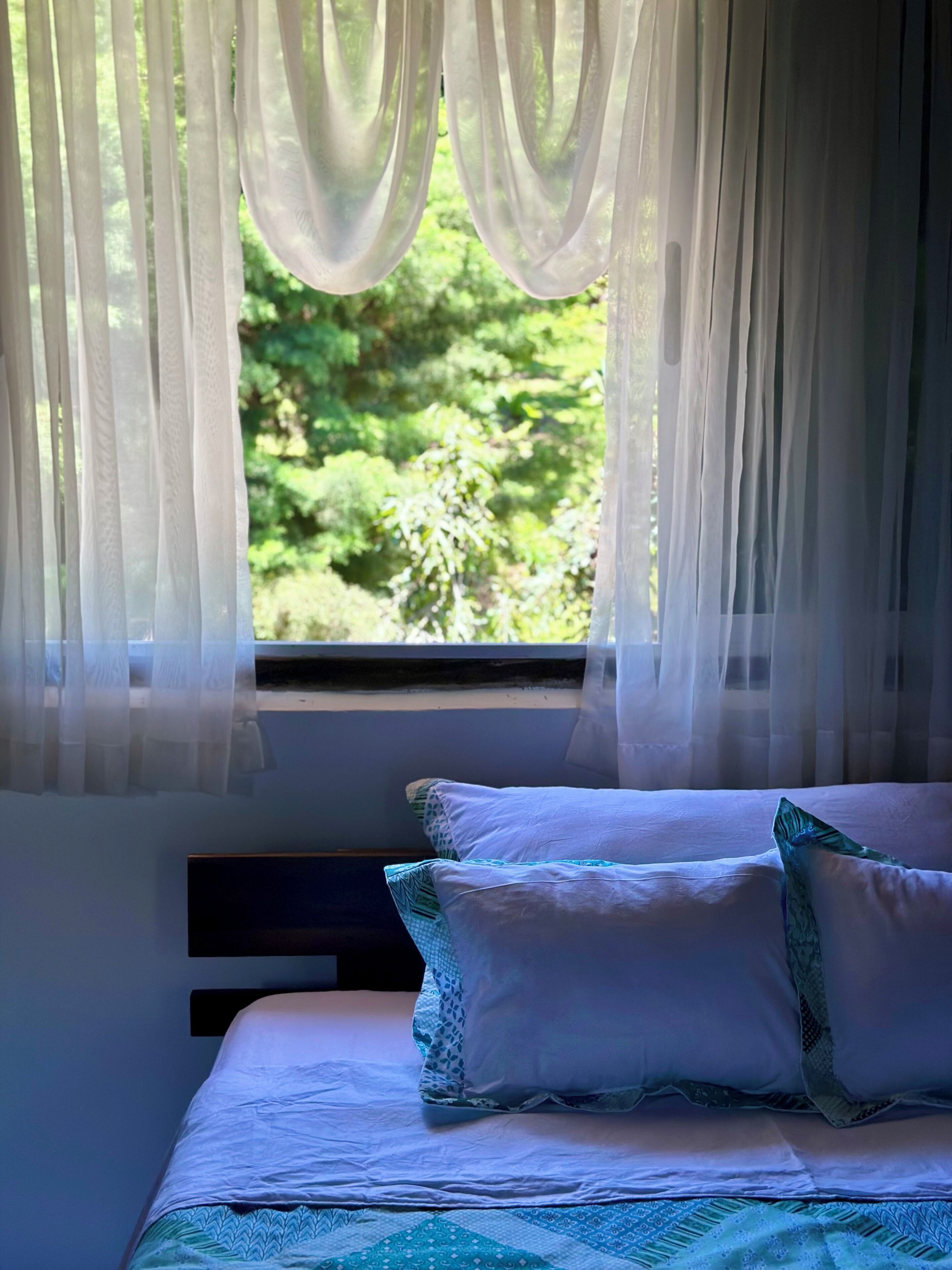 A bed with white bedding and patterned pillows, a window with sheer curtains, and a view of greenery outside.