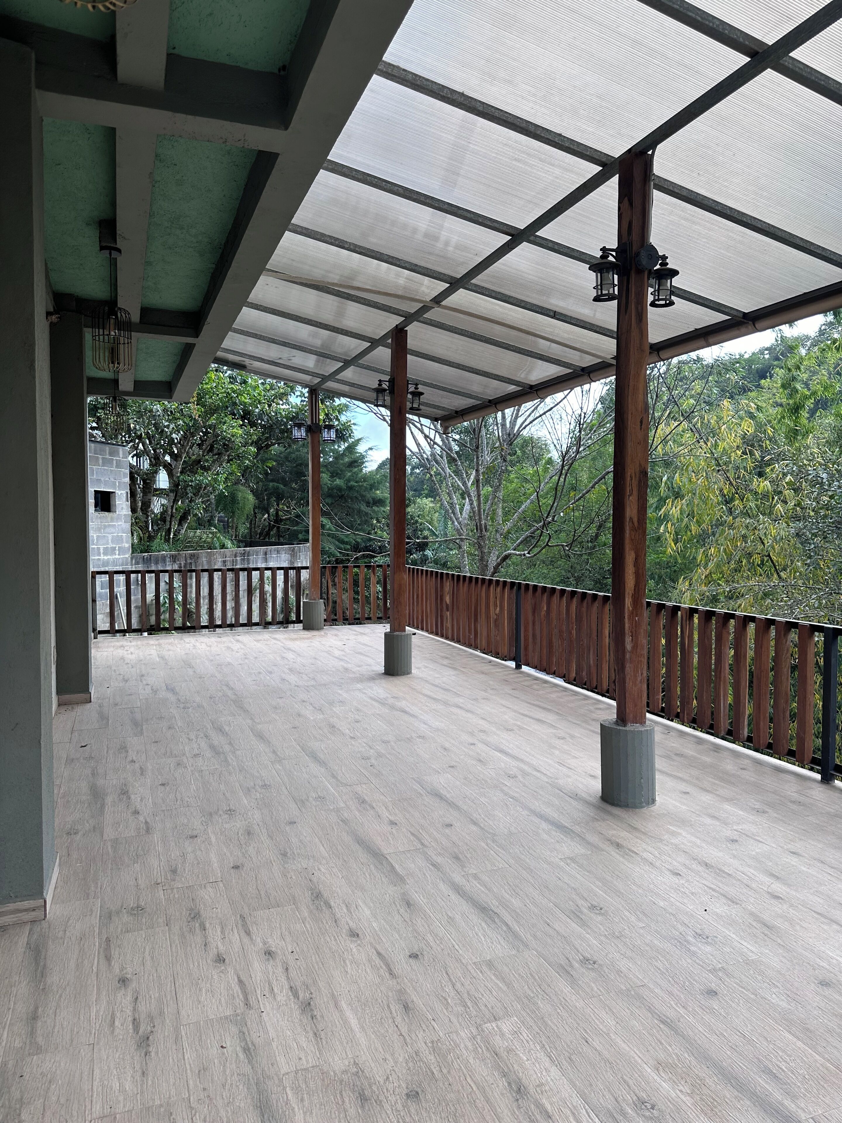 An open wooden deck with a metal railing, supported by wooden pillars, and a translucent roof overhead.