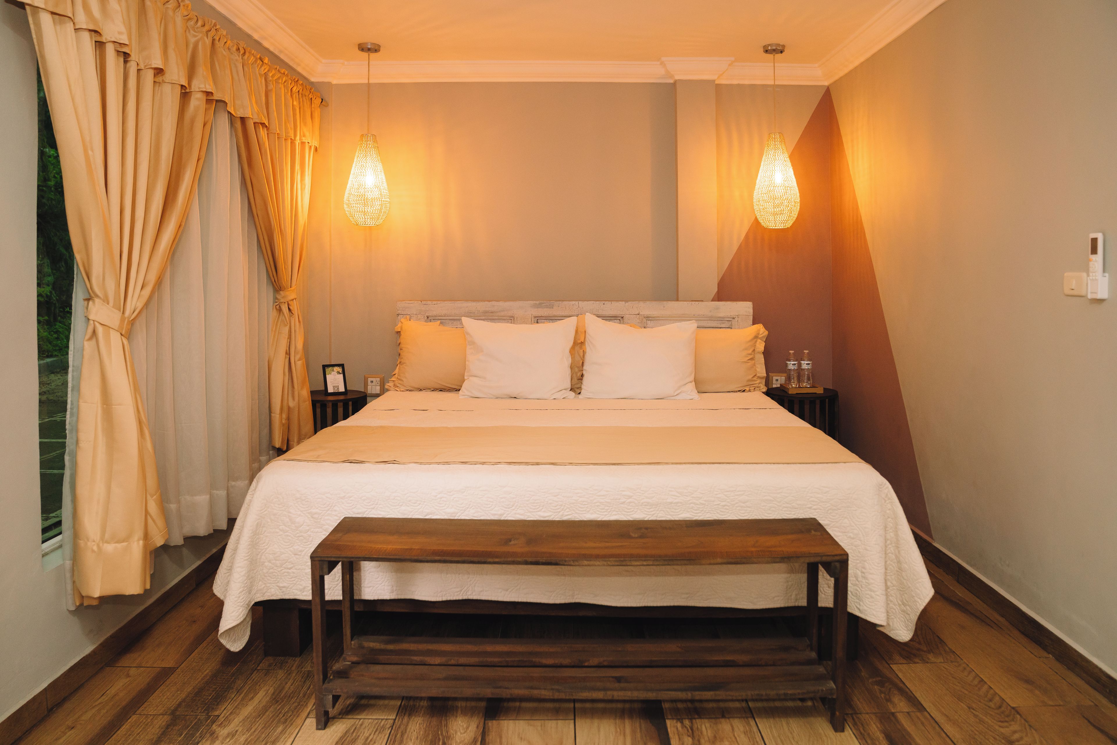 Un dormitorio con una cama de madera, dos mesitas de noche, un banco y una ventana con cortinas.