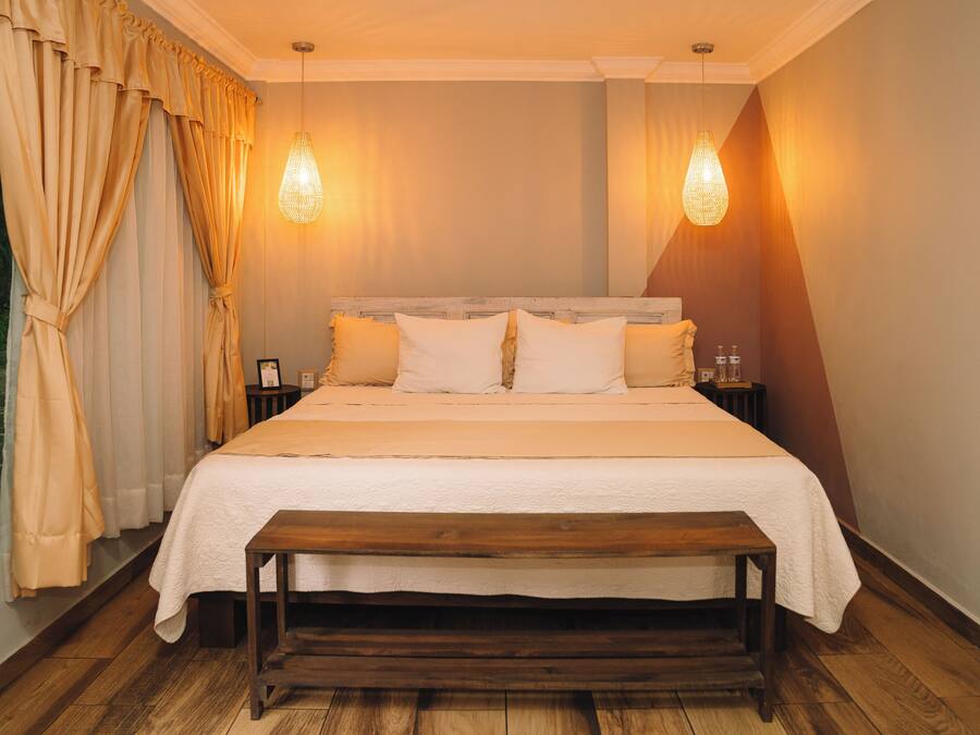Un dormitorio con una cama de madera, dos mesitas de noche, un banco y una ventana con cortinas.