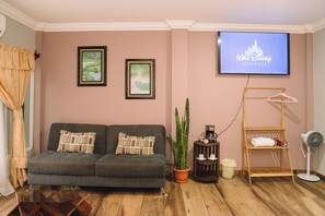 Una sala de estar con un sofá gris, dos cuadros enmarcados, un televisor mostrando un logotipo, una planta en maceta, una cafetera y un ventilador.