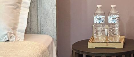 Uma cama com colcha branca, um travesseiro e uma mesa de cabeceira com duas garrafas de água e um suporte.