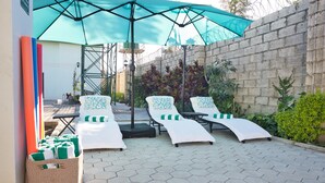 A sunlit outdoor lounge area with hexagonal tiles, three sunbeds with green and white cushions, a large blue umbrella, and a wicker basket filled with folded towels. - F2 - Pool, Inverter, Gym, Pavilion, Wifi, Perfect for Kids - Ibex Meanwood! (Lusaka)