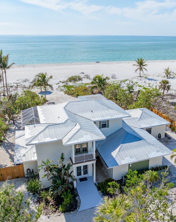 A two-story house with a metal roof, surrounded by a sandy beach and palm trees.