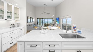 A modern kitchen with white cabinetry, a large island with a sink, and a dining area with a gray sofa and coffee table. - Your Front Yard? The Gulf of Mexico – Blue Gulf 202 Beachfront Condo (Sanibel)