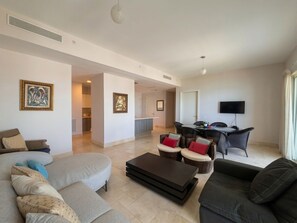 A spacious living area with a sofa, coffee table, and dining table. There is a kitchen in the background and a TV mounted on the wall.