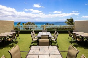 An outdoor patio with tables and chairs, a view of the ocean, and a green lawn.