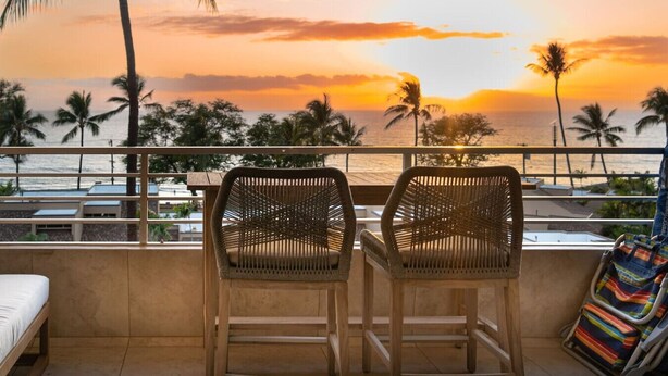 A balcony with two wicker chairs facing a sunset view over the ocean, palm trees, and a railing. - 3 bedrooms 3 baths • Oceans views • Steps to beach (Kihei)