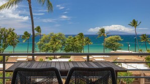 A balcony with a view of the ocean, palm trees, and a wooden table and chairs. - 3 bedrooms 3 baths • Oceans views • Steps to beach (Kihei)