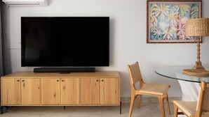 A modern living room with a flat-screen TV on a wooden cabinet, a glass table with chairs, and a tropical-themed painting on the wall. - 3 bedrooms 3 baths • Oceans views • Steps to beach (Kihei)