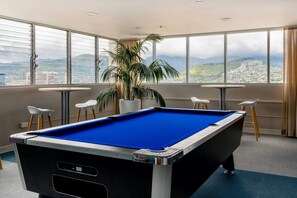 A pool table in a room with large windows offering a city view. - Ocean View•1 BR Corner Condo•Mins to Waikiki Beach (Honolulu)