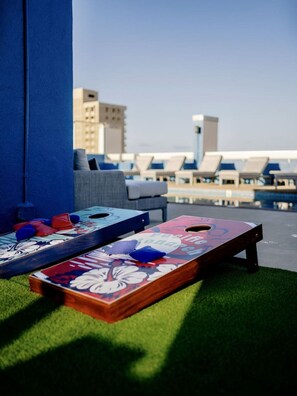 A rooftop terrace with two shuffleboard tables, a seating area with lounge chairs, and a view of buildings in the background. - Ocean View•1 BR Corner Condo•Mins to Waikiki Beach (Honolulu)