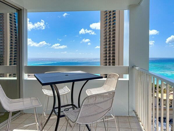 A balcony with a table and chairs, overlooking a clear blue sea and a distant shoreline.