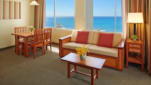 A hotel room with a large window offering a view of the ocean, a sofa, a dining table with chairs, and a small side table with a lamp and plant.