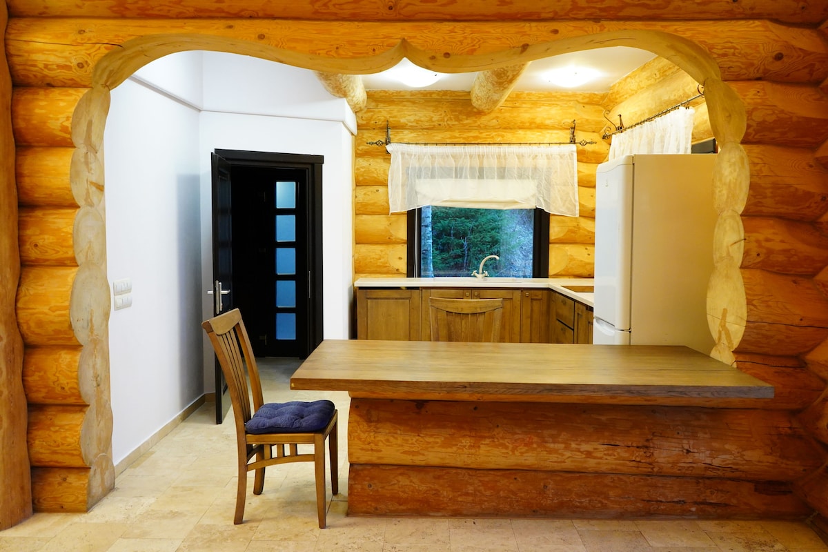 A log cabin interior with a wooden dining table, a chair, a kitchen area with cabinets, and a window with a curtain.