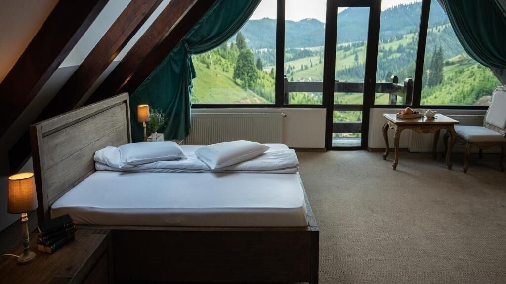 Un dormitorio con cama, mesita de noche, una mesa con silla y vistas a un paisaje verde a través de amplias ventanas.