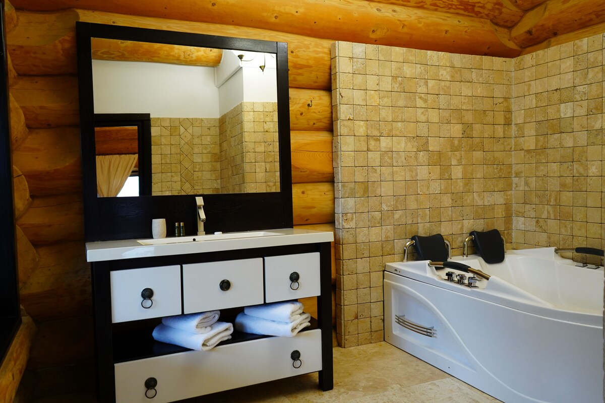 A log cabin bathroom with a wooden ceiling, a large mirror, a white sink with a black countertop, a white bathtub, and tiled walls.