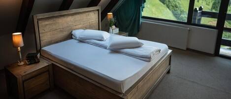 Un dormitorio con una cama de madera, una mesita de noche con una lámpara y vistas a un paisaje verde a través de amplias ventanas.