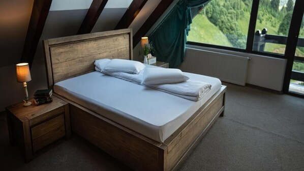 A bedroom with a wooden bed, a nightstand with a lamp, and a view of a green landscape through large windows. - Pensiunea Poiana Izvoarelor (Vatra Dornei)