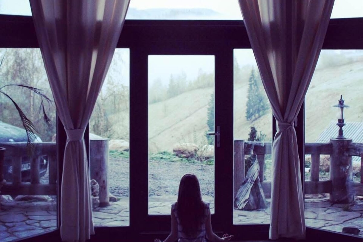 A person meditating in a room with a view of a rural landscape through large windows.