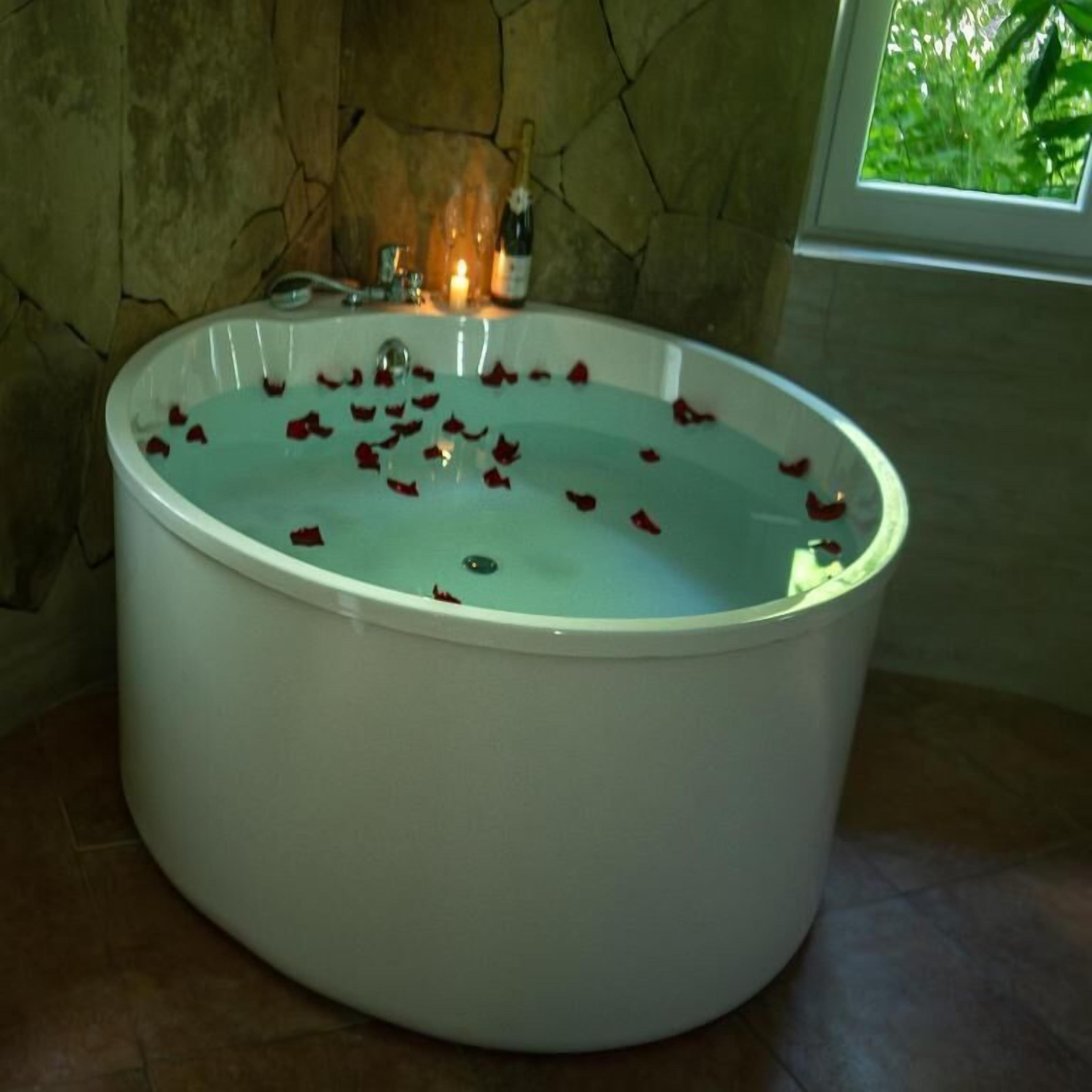 Une grande baignoire ovale remplie d’eau et de pétales de rose, avec une bouteille de champagne et une bougie posées sur le bord.