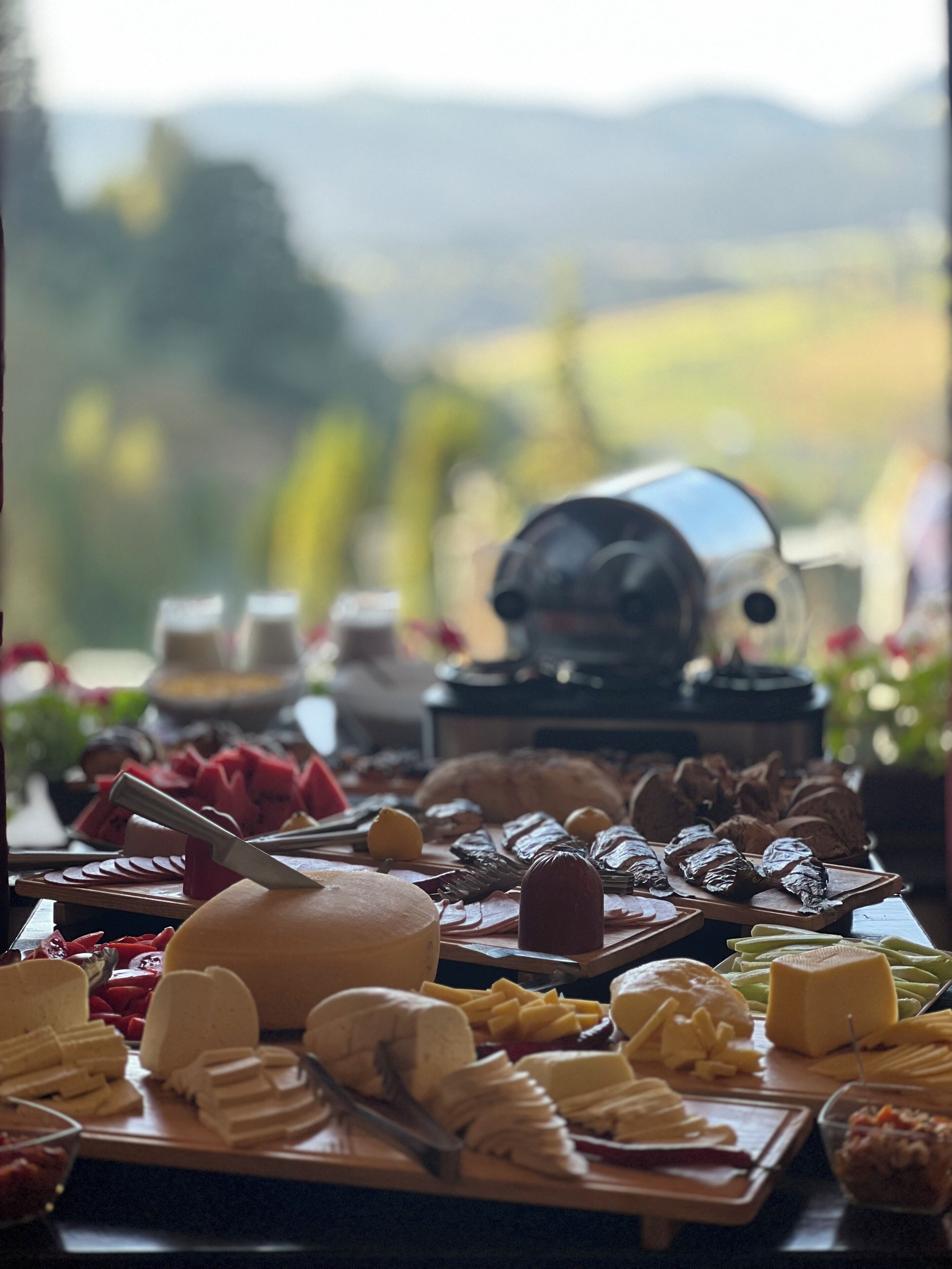 Una tabla de quesos con diferentes tipos de queso y acompañamientos, una cafetera y una vista panorámica al aire libre.