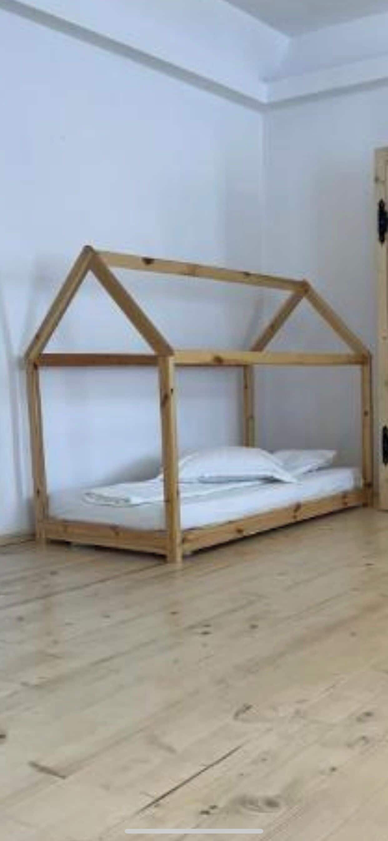 Estructura de cama de madera con forma de casa, ropa de cama blanca y suelo de madera.