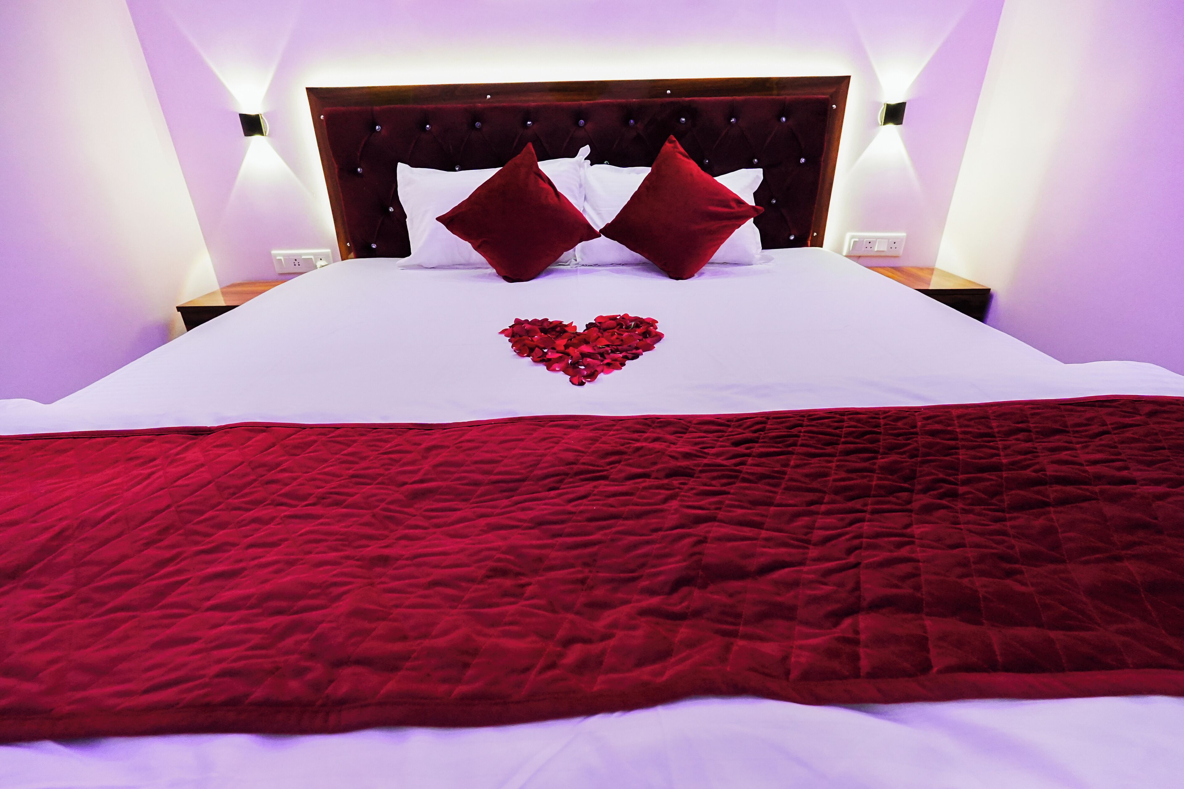 Un letto rifatto con cura, decorato con un’accogliente composizione di petali di rosa a forma di cuore, una coperta trapuntata rossa e due cuscini rossi.