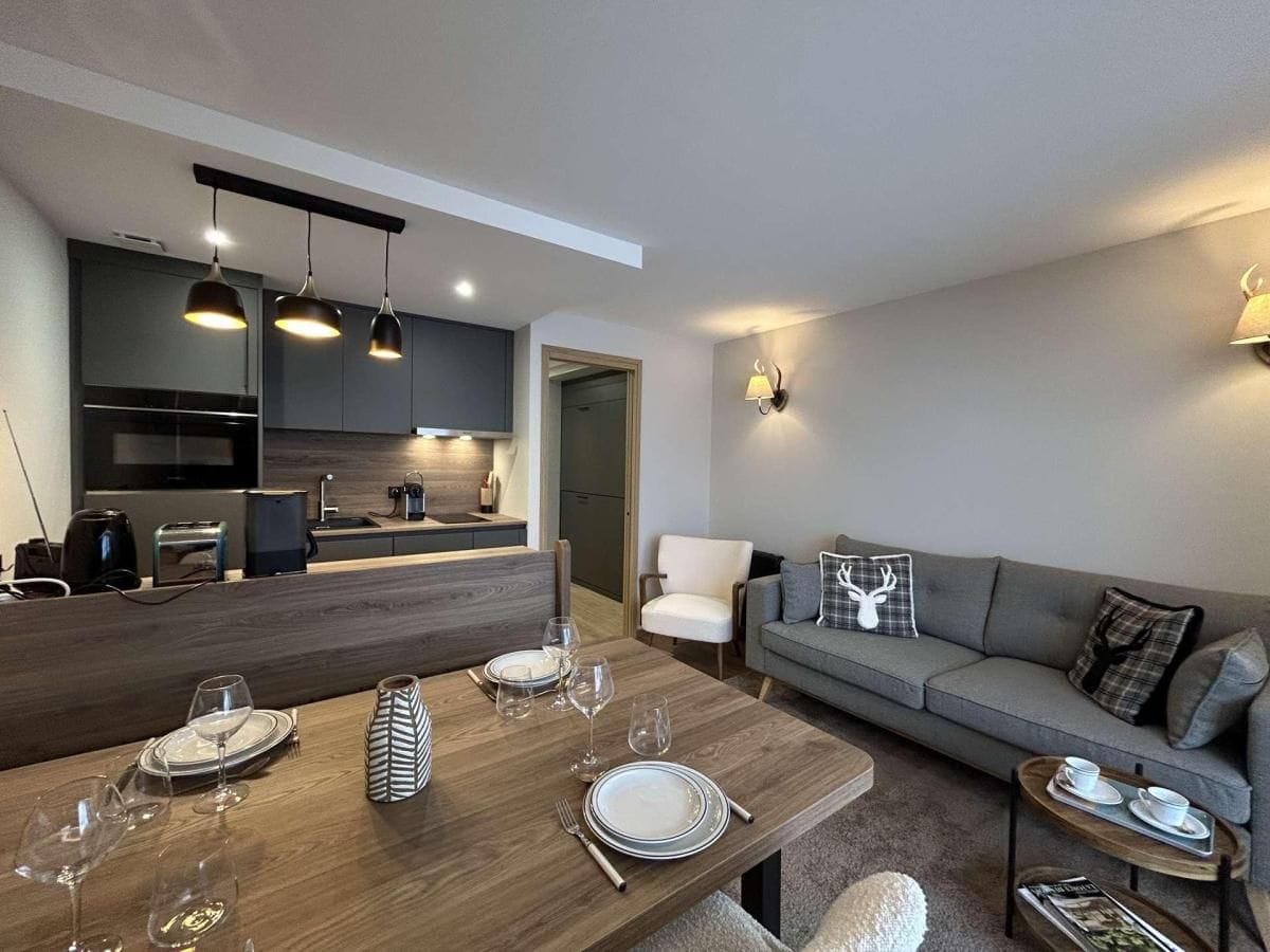 A modern living room with a dining table set for four, a grey sofa, and a kitchen in the background.