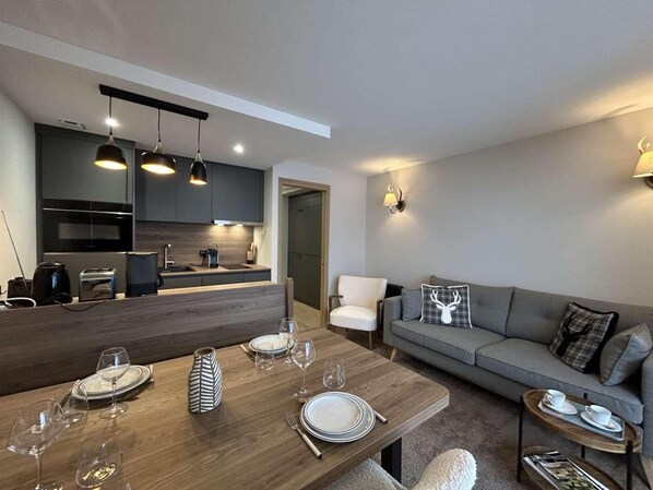 A modern living room with a dining table set for four, a grey sofa, and a kitchen in the background.
