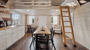 A spacious kitchen with a wooden table and chairs, a wooden ladder, and a view of the outdoors through large windows.