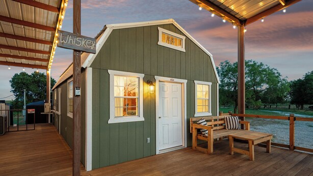 A rustic wooden structure with a 'Whiskey' sign, a covered porch with a bench and table, and a view of trees and sky in the background.