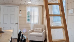 A room with a wooden floor, a white door, a beige armchair with a cushion, a wooden ladder, and a window with blinds.