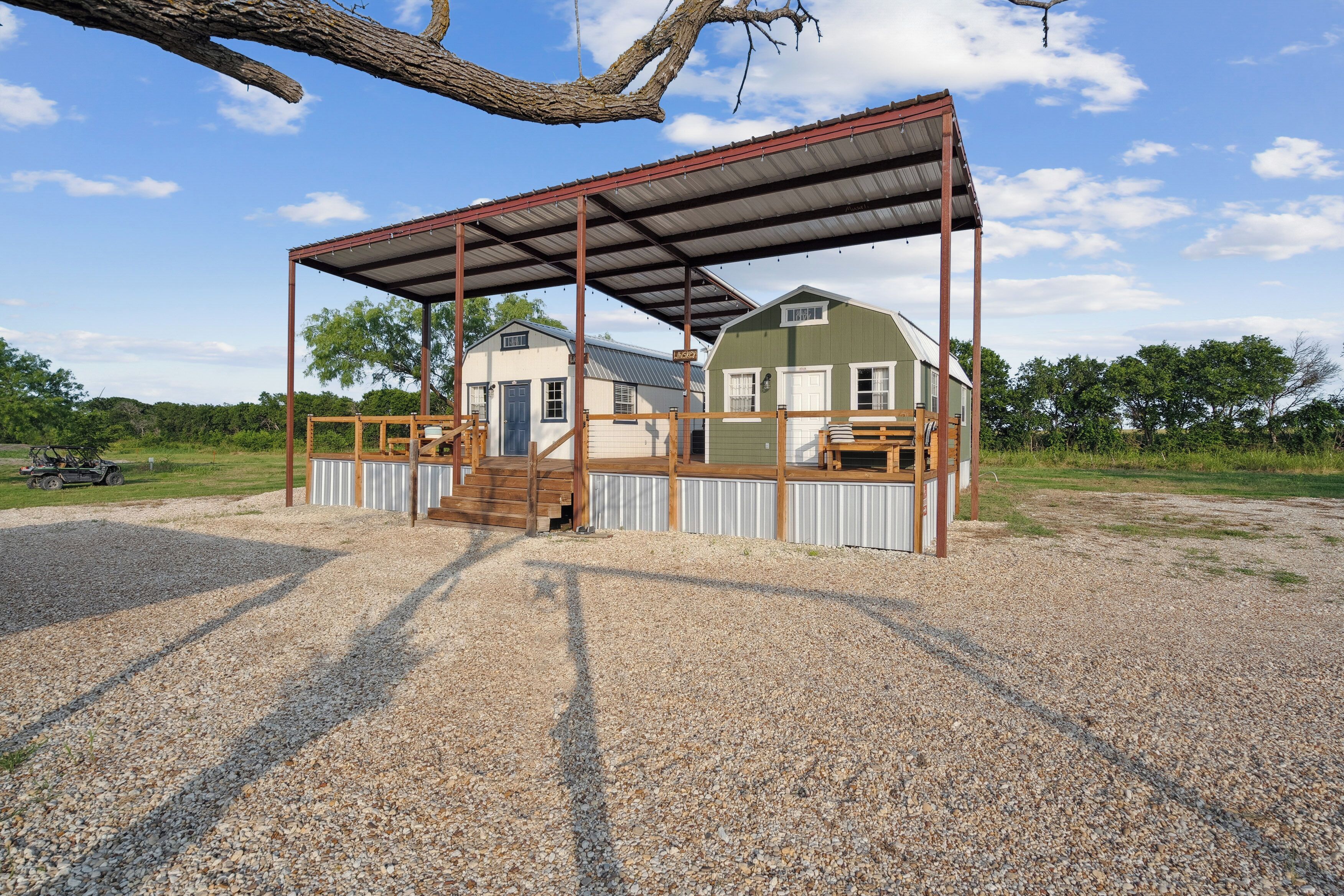A covered outdoor area with a wooden deck, a gravel ground, and a building with a green roof in the background.