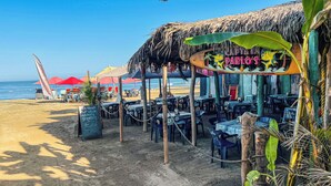 Een restaurant aan het strand met terras, rieten dak en uitzicht op de oceaan.