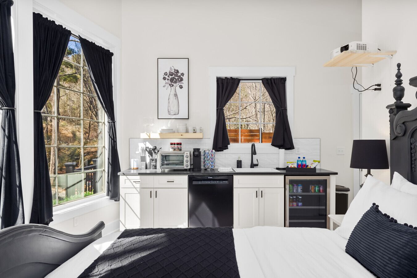 A hotel room with a bed, a desk with a coffee maker, a dishwasher, and a view of trees through large windows.