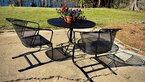 A round metal table with two chairs, a potted plant with purple flowers, and a view of a lake and trees in the background. - Sleeps 10. Bayou Front Home close to ULM, Sports Complexes, Airport and I-20. (Monroe)
