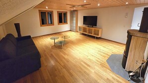 Ein Wohnzimmer mit geneigter Decke, Holzboden, einer schwarzen Couch, einem Fernseher auf einem Holzschrank und einem Glastisch.