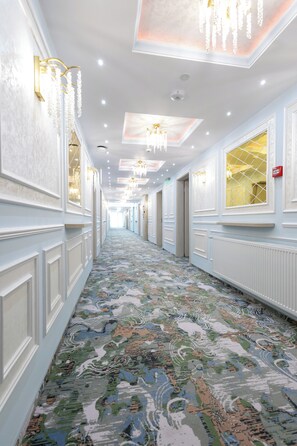 A long corridor with ornate wall panels, chandeliers, and patterned carpet.