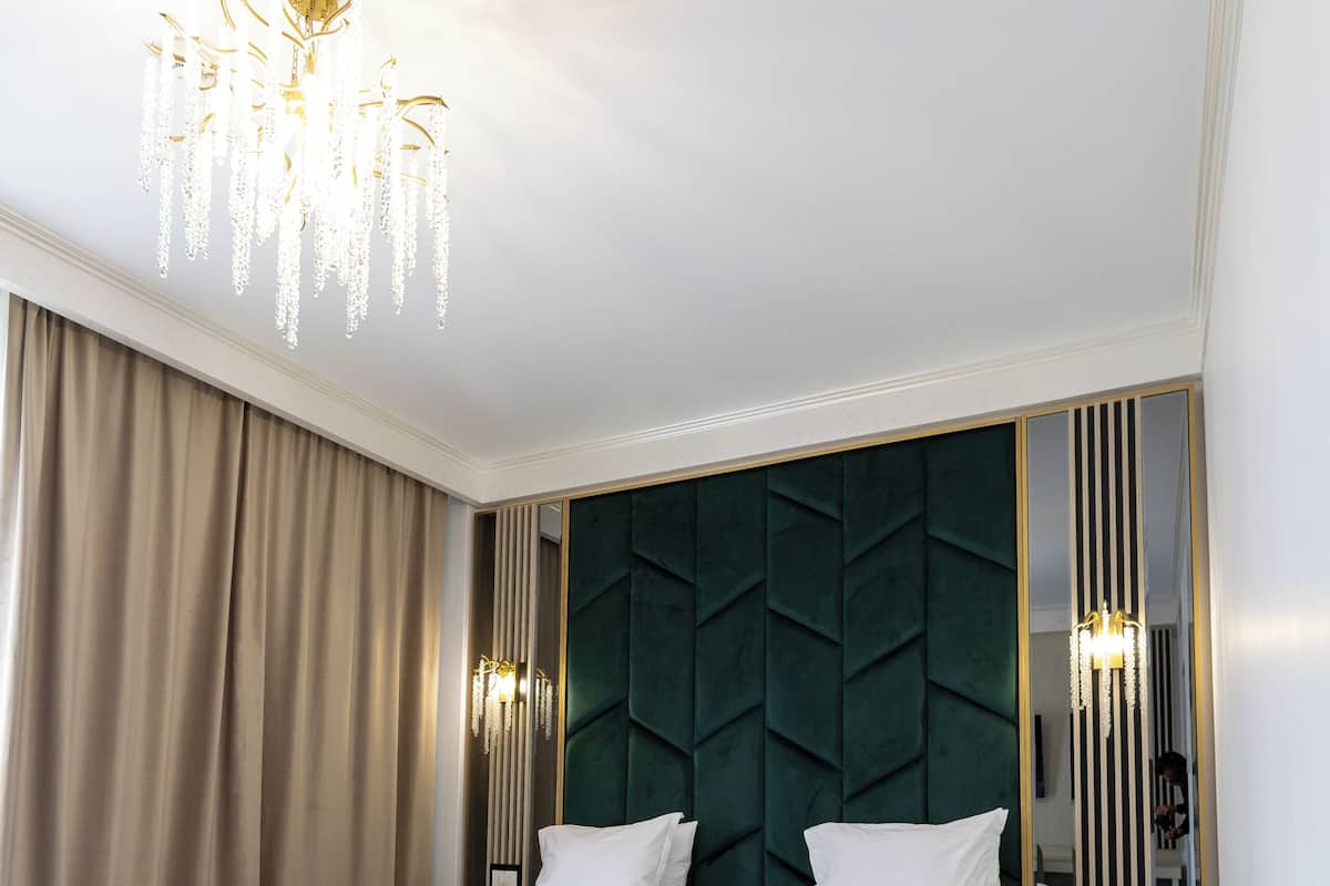 A hotel room with two beds, a chandelier, a green upholstered headboard, and a patterned carpet.